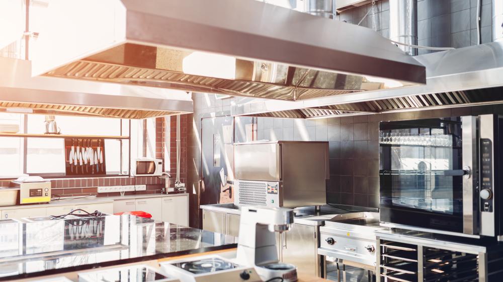 A sunny industrial kitchen with equipment, a hood over a range, and ovens. 