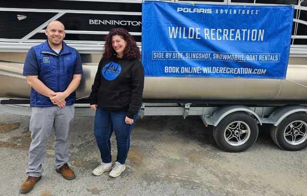 A woman and man stand in front of a pontoon boat with a banner that says the company name, "Wilde Recreation."