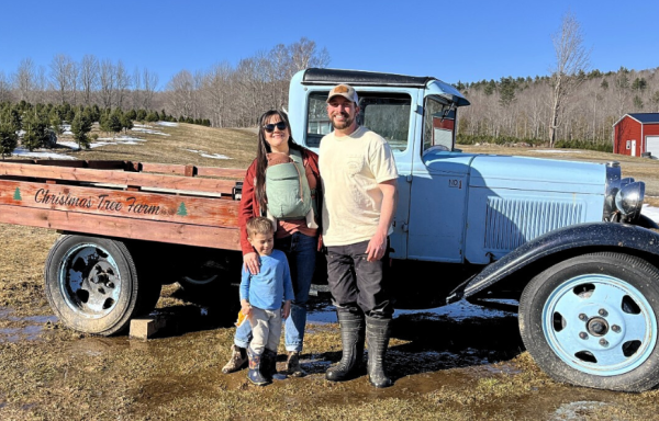 Molly and Jesse of Piper Mountain Christmas Trees stand in front of a vintage blue pickup truck with their two children. In the background there is a red barn and a distant hill. 
