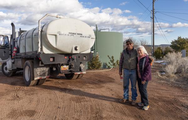 Two people standing in front of a water tank and water truck