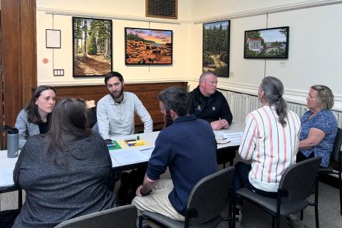 Seven people are seated around a rectangular table talking to each other. There is artwork on the walls in the background.