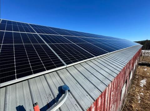 The image shows solar panels on the roof of a red barn under a blue sky.