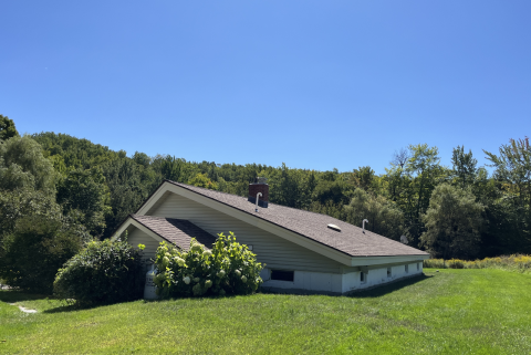 A home is set into a hillside with the roof and a few windows visible. It is surrounded by green lawn, with trees in the background and a bright blue sky overhead.