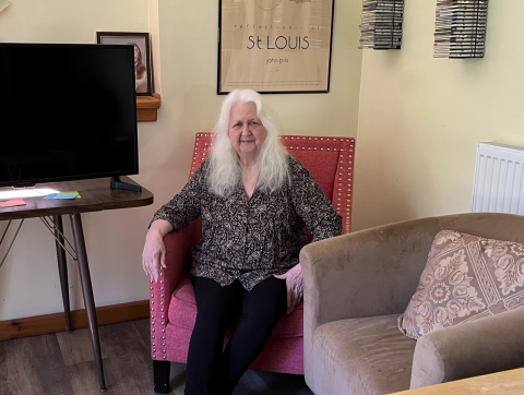 A woman is shown seated in an arm chair in a living room with artwork on the walls and a tv to one side.