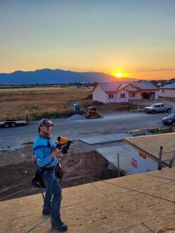 Cody standing on an unfinished rooftop with a nail gun. Incomplete house in background has a sun setting behind a Northern Utah mountain.