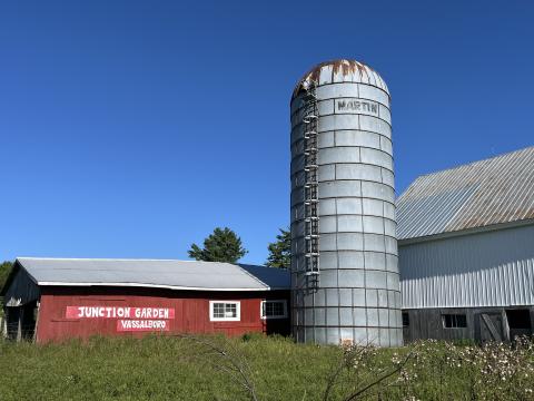 A low red farm building says "Junction Garden Vassalboro." There is a silver silo and a taller barn to the right, and blue sky overhead. 