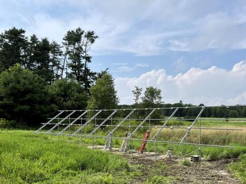 The structural support for the Junction Garden solar array is shown in a green field with trees in the background. The installation is under construction in the photo.