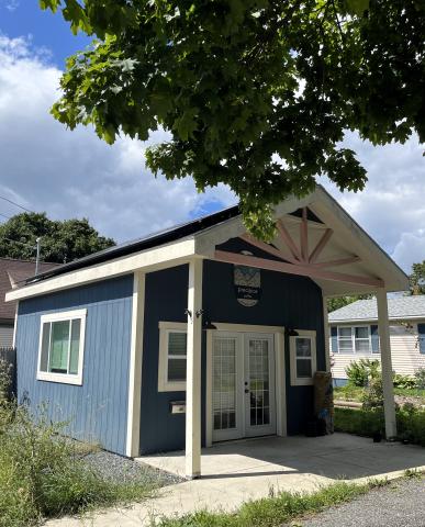 The company roasts the coffee in a small shop that is blue with white trim and has a porch. The solar panels are on the rood. It is shown on a sunny day with a leafy tree overhead. 