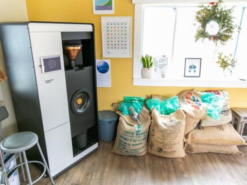 The Bellweather Roaster is about the size of a domestic refrigerator. It is shown in a yellow room near a window with bags of coffee stacked nearby.