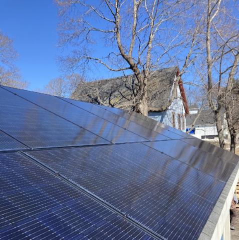 Solar panels cover the entire roof of the Precipice Coffee Ellsworth location, a small building in a residential neighborhood. A nearby house and leafless tree can be seen in the background.