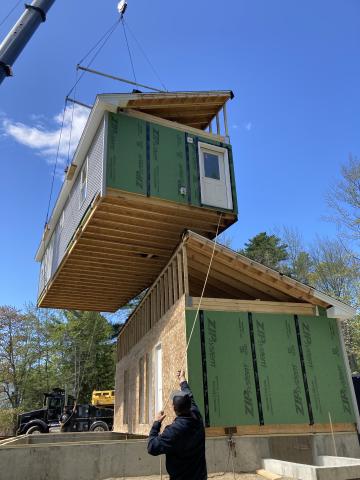 A modular home is shown being assembled using a crane. One segment of the house is already on the foundation and another segment is being lowered into place. 