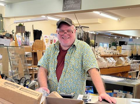 A man stands at the checkout area of the store, with bright lights overhead and aisles full of goods behind him.