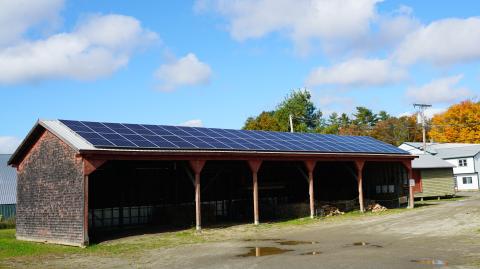 A long, low pole bard behind the Marsh River Co-op contains firewood and has solar panels across the roof. There are residences in the background and fall foliage visible.