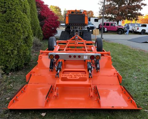 The club's orange "Trail Paver II" trail dragger is shown from behind, attached to a grooming machine, on a lawn near a parking lot. 