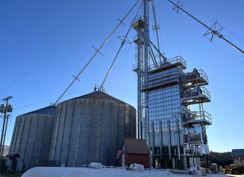 The grain dryer and adjacent silos are shown against a blue sky.
