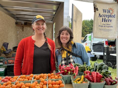 Two women stand at a farmers' market stall with bins of produce in front of them and an open truck behind them.