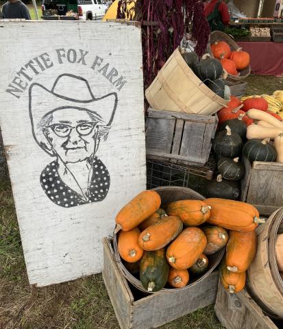 A sign that says "Nettie Fox Farm" is leaned up against a tent pole and surrounded by bins of fall produce.