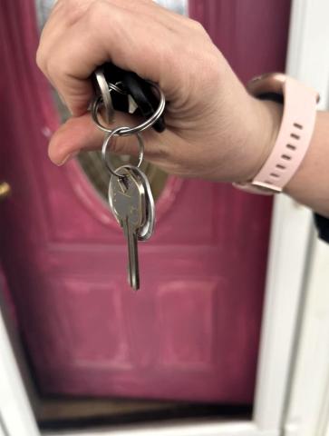 A hand is shown holding a set of keys in front of a magenta house door that is ajar.