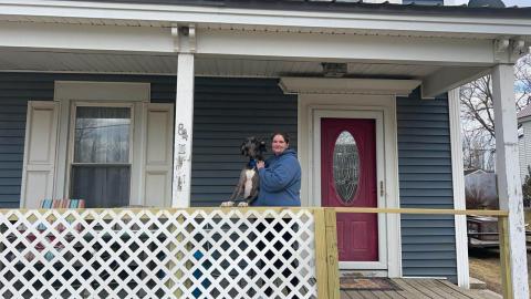 A woman stands on the front porch of a grey house with white trim with a large dog at her side. 