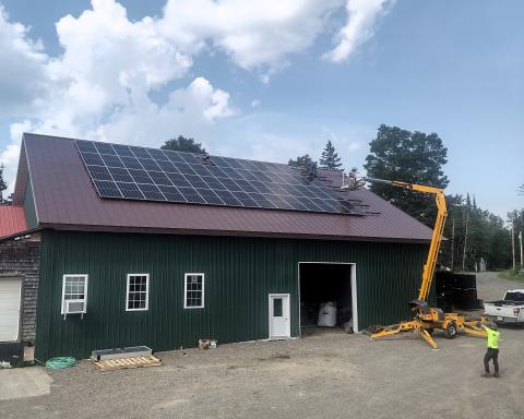 The image shows the solar panels arranged on the roof of the Wood Prairie Family Farm warehouse with a crane completing installation.