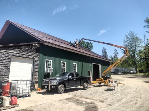 The image shows the rooftop solar panels being installed on the Wood Prairie Family Farm building under a sunny sky.