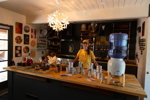 A woman in a yellow shirt and hat stands behind a kitchen island in the lodge at a guest accommodation. A display of locally-produced products rests in front of her.