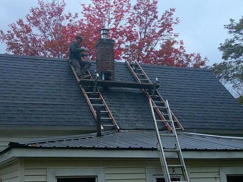 A man works on a chimney on the roof of a house with wooden scaffolding around and fall foliage in the background.