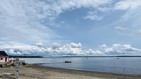 The image shows an empty beach in the foreground with a receding tide. There is blue sky and clouds overhead.