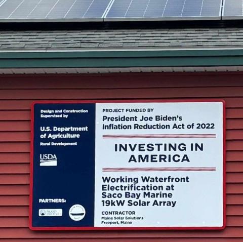 A red, white, and blue sign says "Investing in America: Working Waterfront Electrification at Saco Bay Marine." The sign is mounted on a building with red siding. The edge of black solar panels is just visible on the roof overhead.