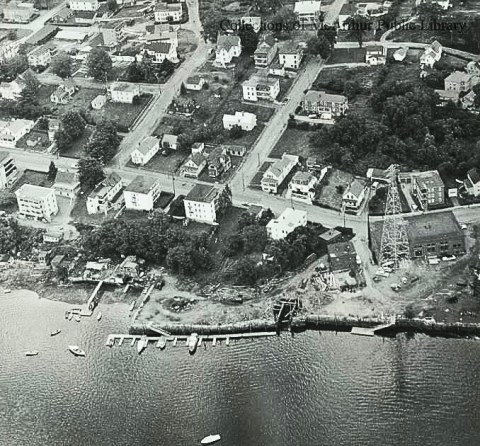 An old black and white photo shows Rumery's Boat Yard from the air in 1955. Some of the old electric plant infrastructure can still be seen in the photo. Houses and streets surround the boat yard, and the Saco River is visible at the front.