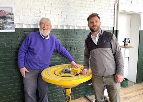 Two men pose on either side of a round, yellow, waist-high device. They are indoors with a cement wall behind them. The device is a solar-powered water quality testing buoy. 