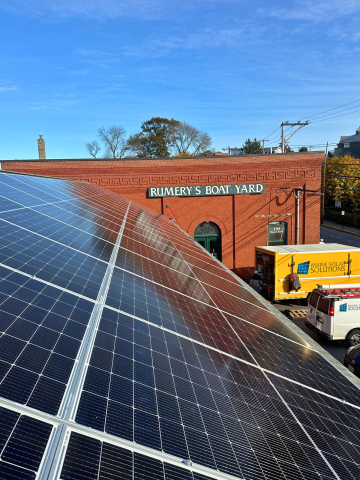 The photo shows solar panels mounted on a long, narrow roof as seen from the roof. Another part of the building in the back ground bears a sign, "Rumery's Boat Yard." Solar company trucks are visible in the background. There is a blue sky and leafless trees in the far background.