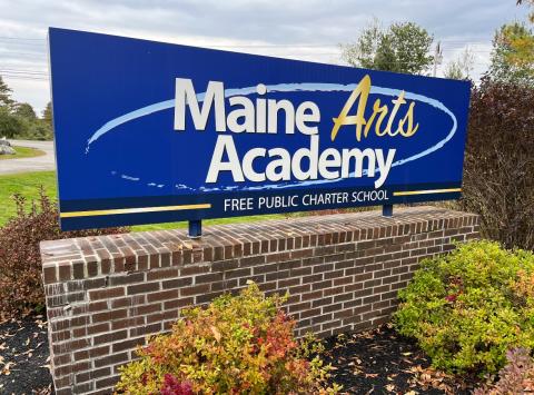 The Maine Arts Academy sign is mounted on a brick base with shrubs and other greenery surrounding it.
