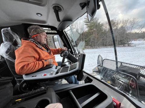 A man in an orange jacket drives a snow grooming machine through a snowy field.