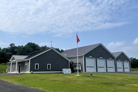 The Corinna Fire Department is pictured. It has grey siding and white trim, with offices on the left and 5 bays on the right. 
