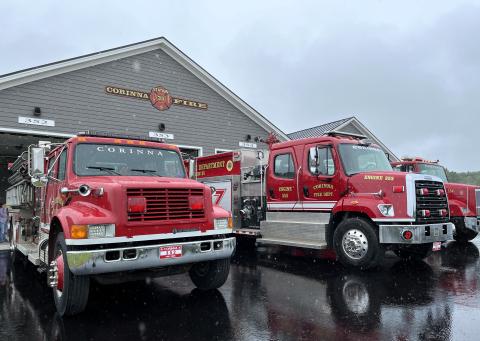 Two fire trucks are pictured parked in front of the Corinna Fire Department on a rainy day. The paved driveway is wet with rain and the red trucks are reflected. The station at the rear is grey.