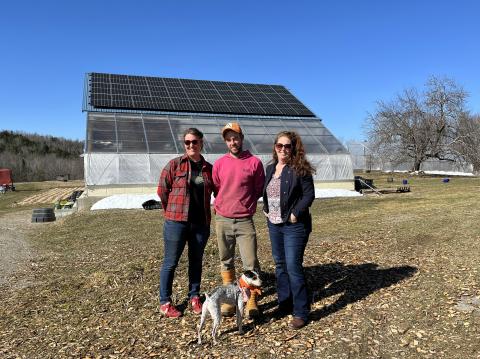 Three people and a small dog pose in front of a barn with solar panels on the roof. The ground and trees are bare and there is a bright blue sky overhead.