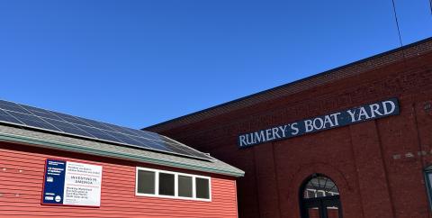 A large red building has a sign on the right that reads "Rumery's Boay Yard" and a smaller sign on the left that is illegible. There is a solar array on the roof of the left roof. 
