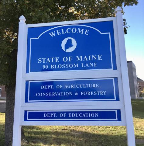 A blue sign reads "Welcome State of Maine Department of Agriculture, Conservation and Forestry." There is a tree overhead and a brick building and lawn behind the sign.