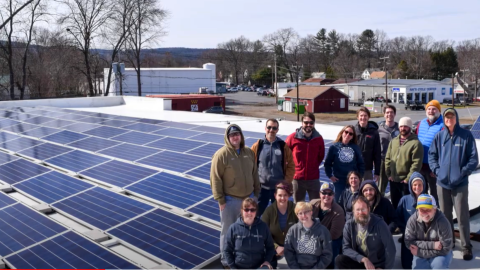 Members of the Real Pickle Co-op stand on the roof of the building next to the new solar panels on a sunny day. Many are wearing coats and it looks chilly outside.