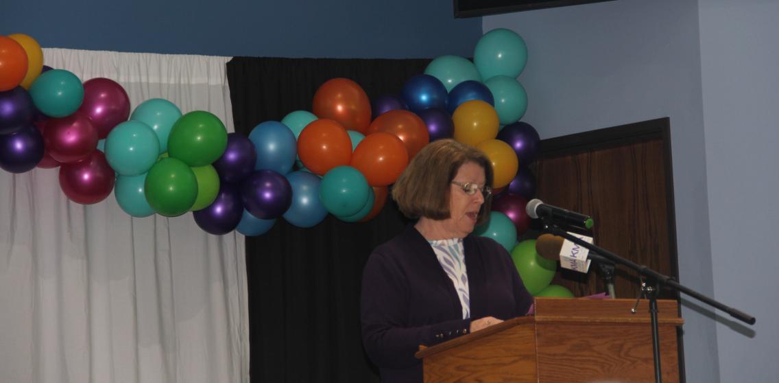 A woman speaks from a podium on a stage decorated with balloons.