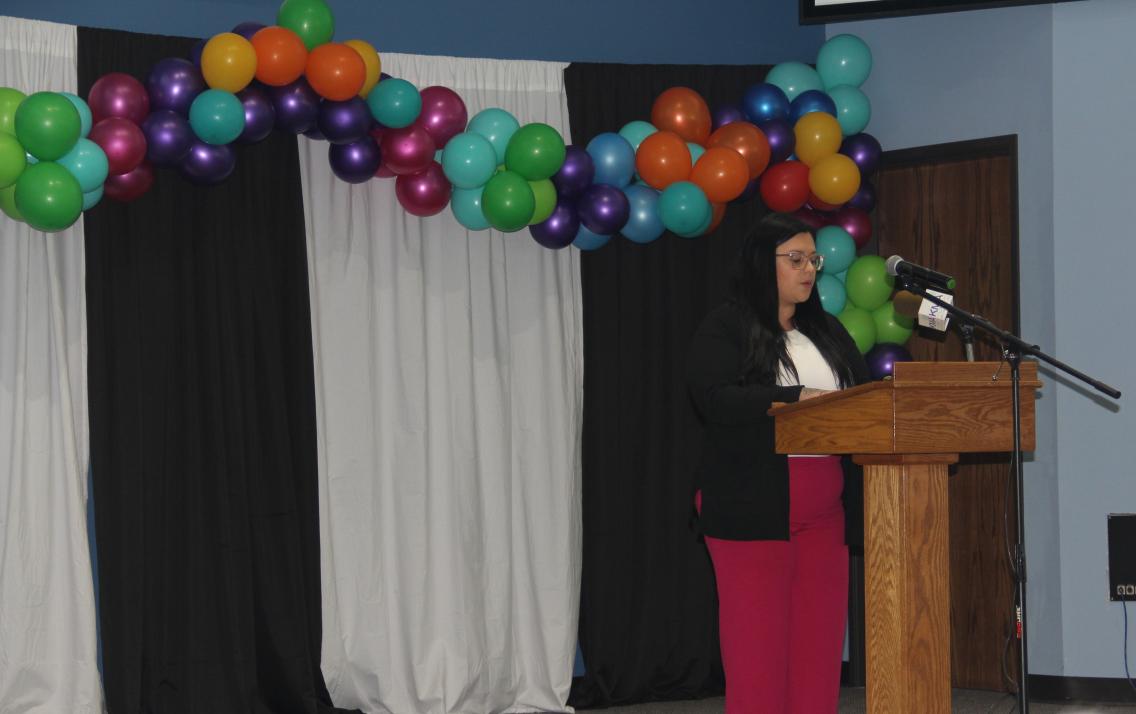 A woman stands at a podium with balloons behind her.