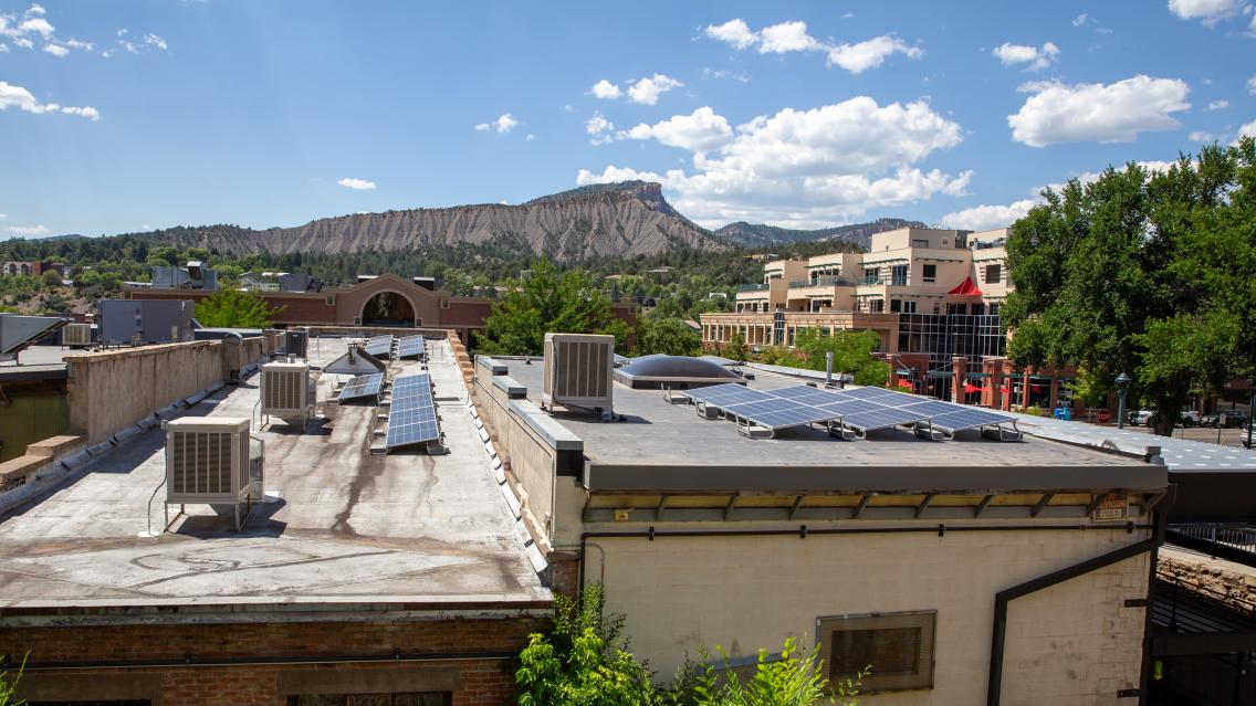 Solar panels are fixed to the roof of a gym. Colorado mountains are seen in the background under a blue sky with a few white clouds.