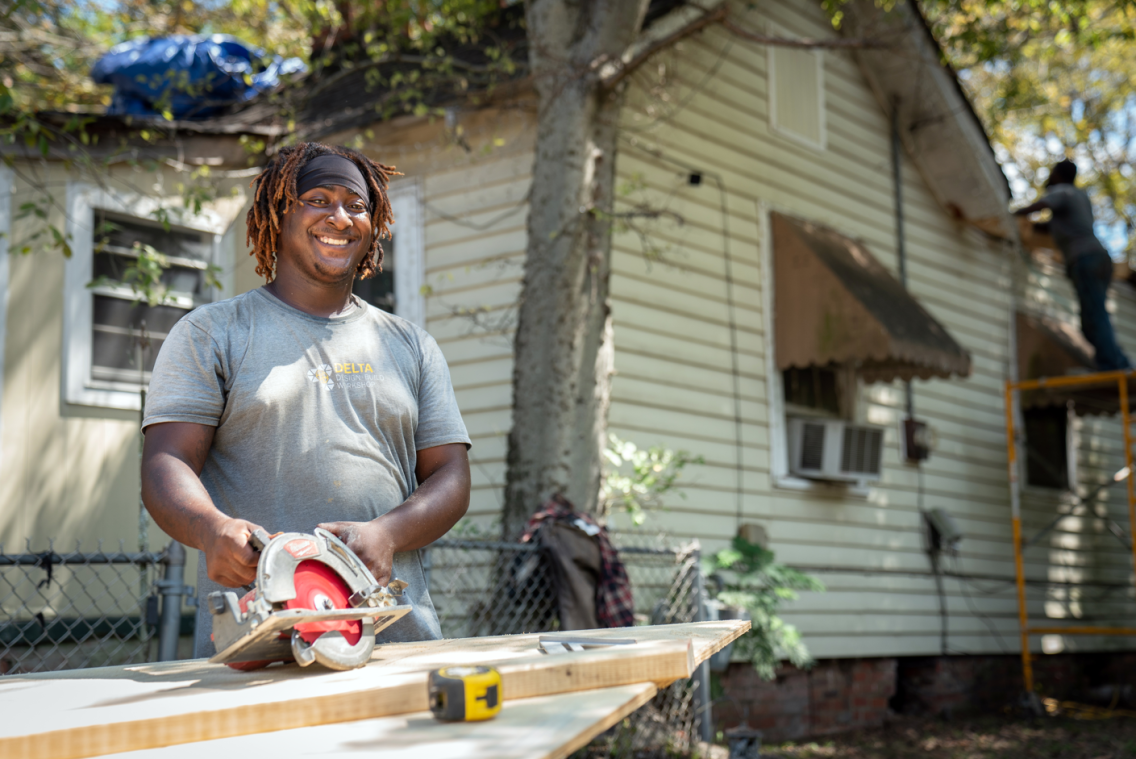 A smiling man standing in front of a yellow house. He is holding a circular saw resting on a piece of wood. Another man works in the background.