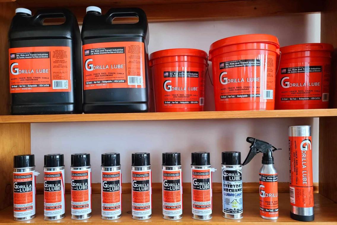 Gorilla Lube products are shown lined up on a shelf. The labels are bright orange. There are jugs, buckets, and spray cans.