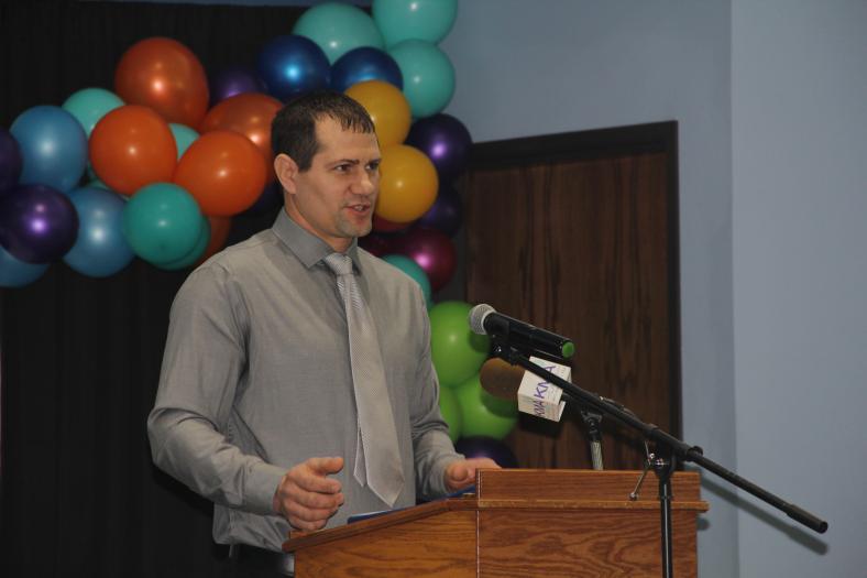 A man stands and speaks at a podium on a stage decorated with balloons.