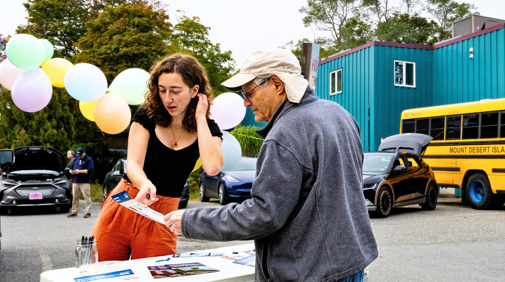 The Climate Fellow described in the story is pictured at an outdoor table interacting with a community member who is reading a pamphlet she shows him. There is a blue building, a Mount Desert Island School bus, and trees in the background. The event pictured is in a parking lot, with balloons in the near background.