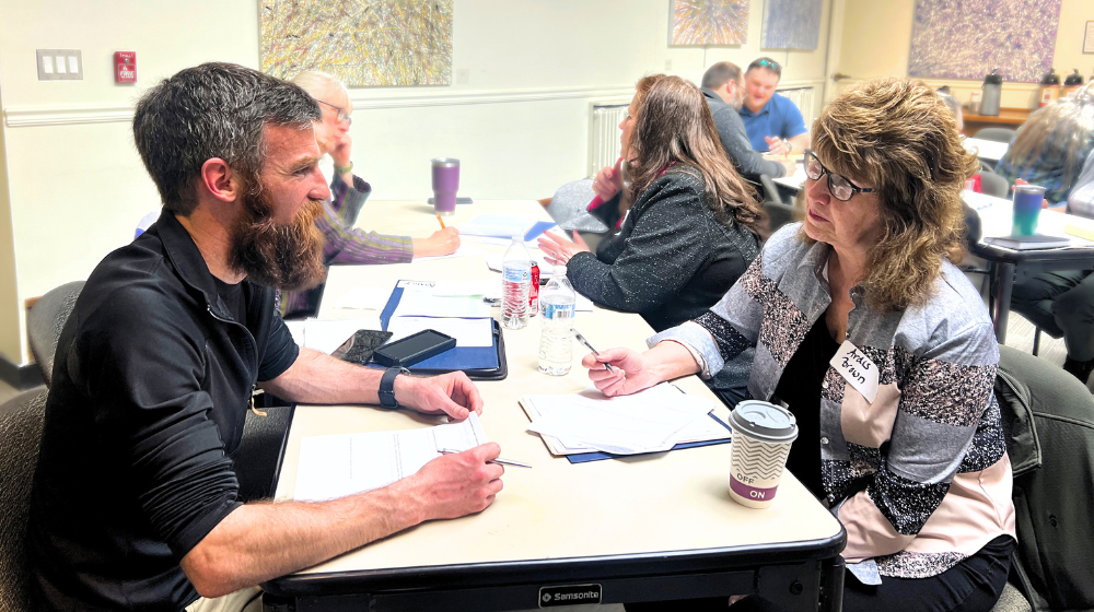 A man and a woman are pictured seated at a table, talking and taking notes. Other people work at the same table and other tables in the background. There is abstract artwork on the walls in the background.