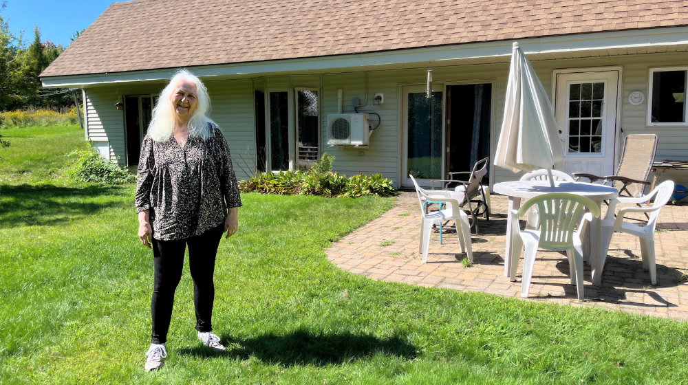 A woman stands before a cream colored house set into a hillside, with a brick patio and white patio furniture arranged. There is a green lawn in the foreground and trees in the background in bright sunshine.