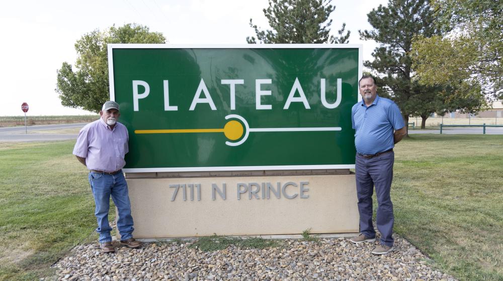 Two men stand next to a green sign that says Plateau